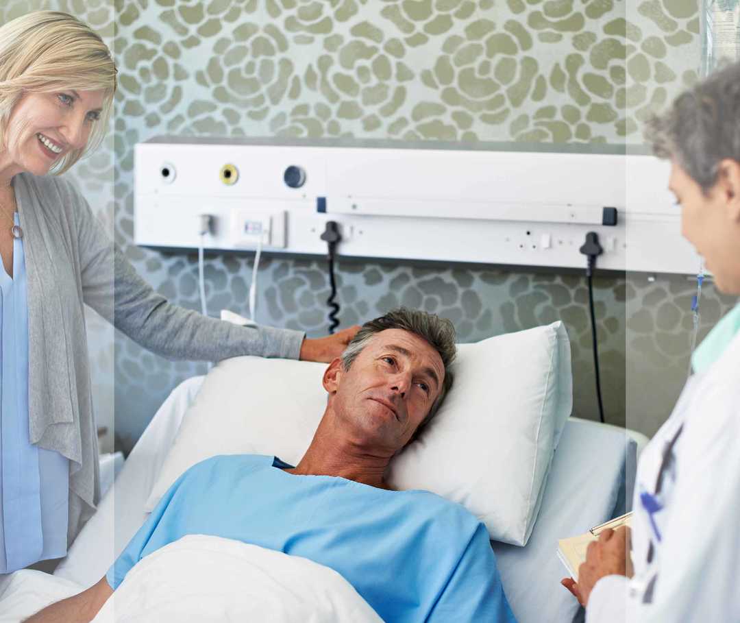 Mann im Krankenhausbett und seine Frau unterhalten sich mit einer Ärztin.