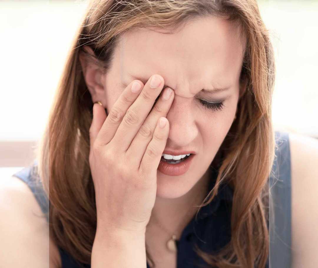 Frau fasst sich ins schmerzverzerrte Gesicht und bedeckt mit der Hand ein Auge.