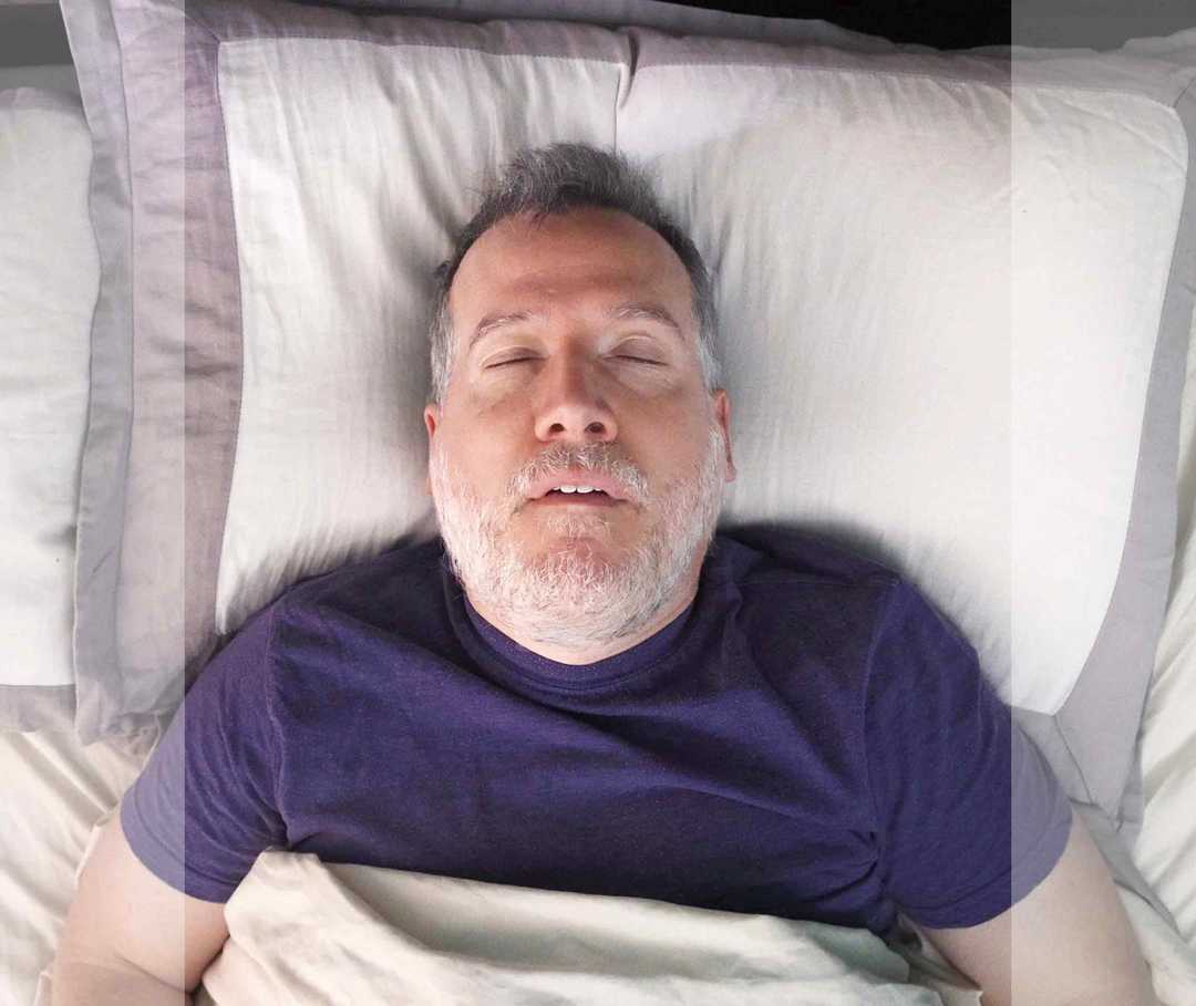 Ein älterer Mann liegt im Bett und schläft. Man sieht seinen Oberkörper und sein bärtiges Gesicht.