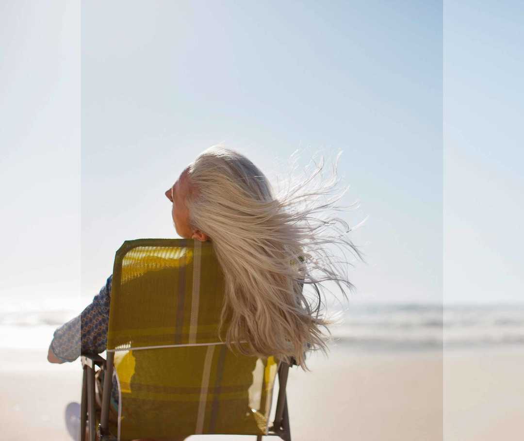 Langhaarige blonde Frau sitzt mit dem Rücken zum Betrachter in einem Campingstuhl am Strand und ihr Haar weht im Wind.