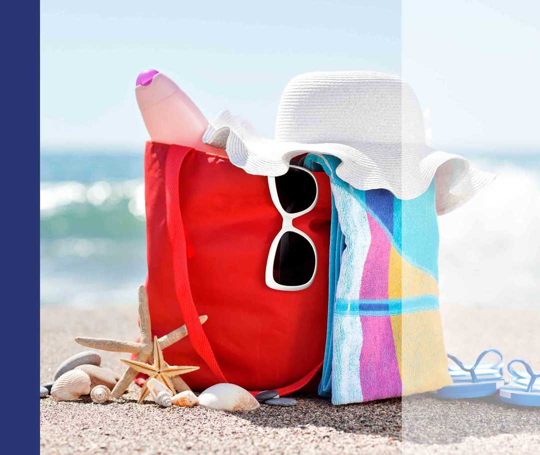 Eine rote Tasche steht am Strand. Enthalten sind Strohhut, ein buntes Handtuch und eine Sonnenbrille.